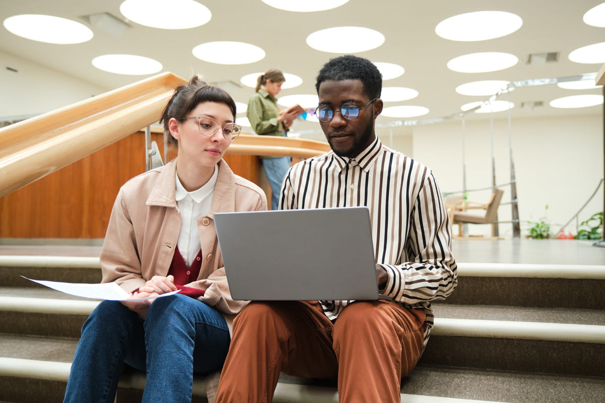 Zwei junge Erwachsene sitzen nebeneinander auf einer Treppe und schauen auf einen Laptop.